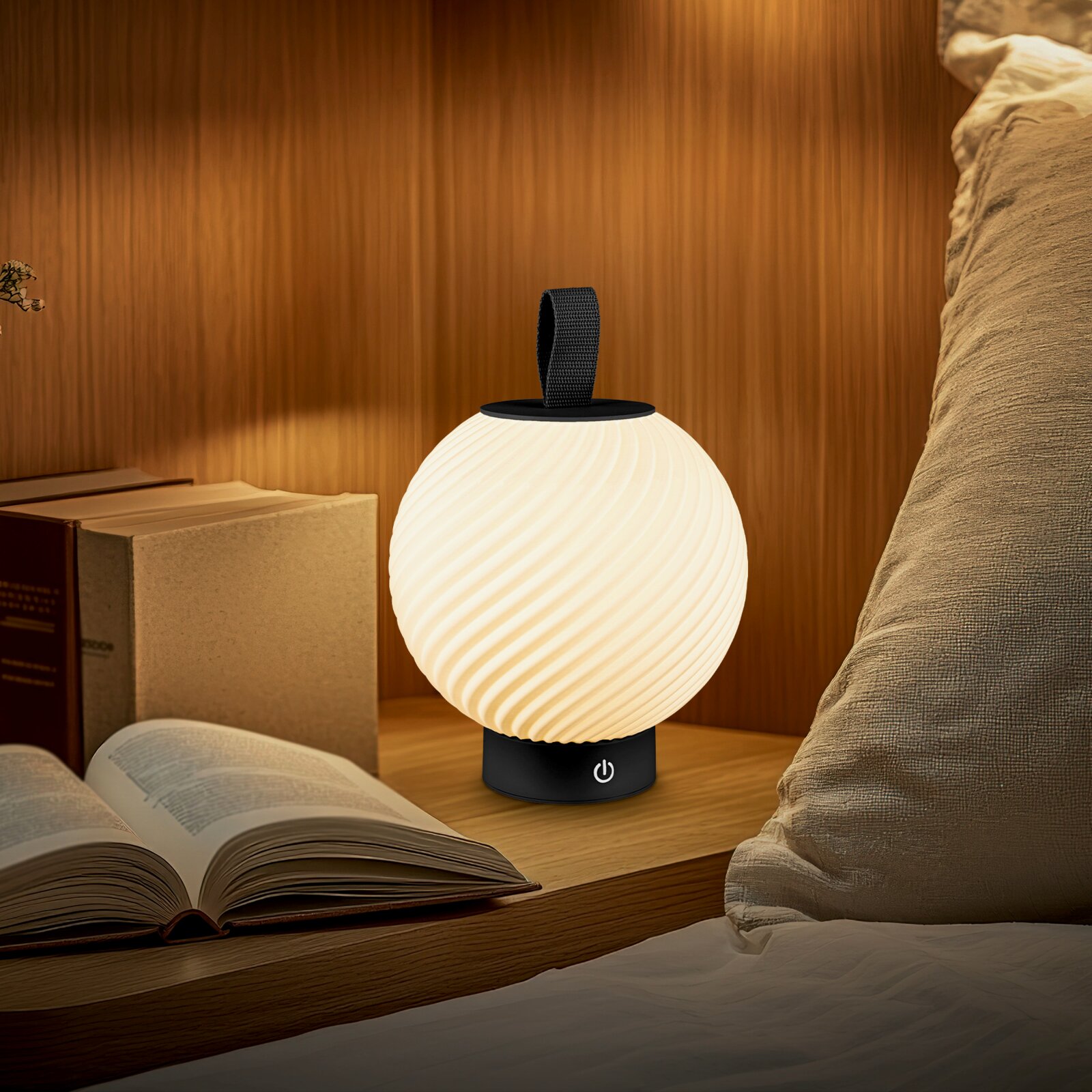 Leuchtende, kugelförmige LED-Tischlampe mit geriffeltem weißen Schirm, schwarzem Sockel mit Power-Symbol und Trageschlaufe, auf einem Nachttisch neben Büchern und Bettkissen – фронтale Perspektive auf Augenhöhe