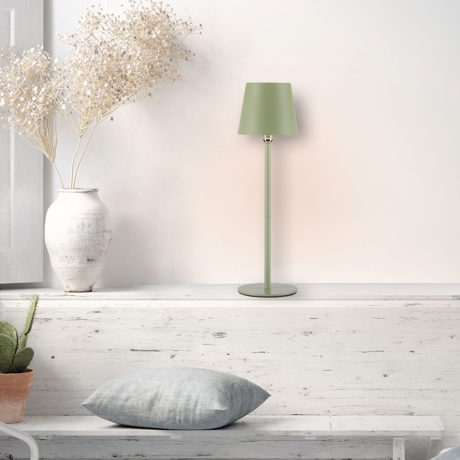 Grüne Akku-Tischleuchte BOTERIA auf einem weißen Regal, seitliche Perspektive, neben einer weißen Vase mit Trockenblumen.