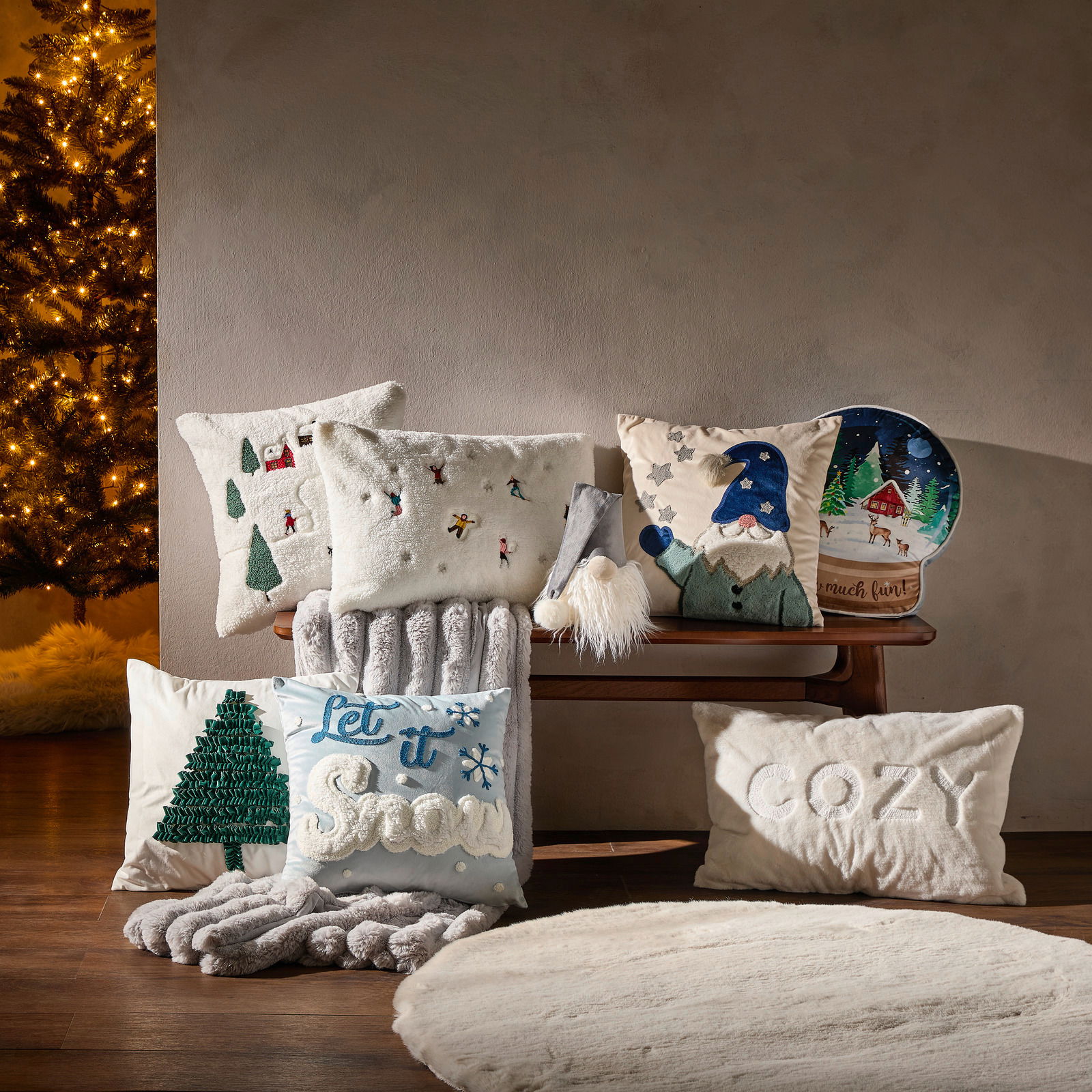 Verschiedene dekorative Kissenhüllen mit winterlichen und weihnachtlichen Motiven, auf einer Holzbank arrangiert, mit einem beleuchteten Weihnachtsbaum im Hintergrund. Perspektive von vorne.
