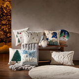 Verschiedene dekorative Kissenhüllen mit winterlichen und weihnachtlichen Motiven, auf einer Holzbank arrangiert, mit einem beleuchteten Weihnachtsbaum im Hintergrund. Perspektive von vorne.