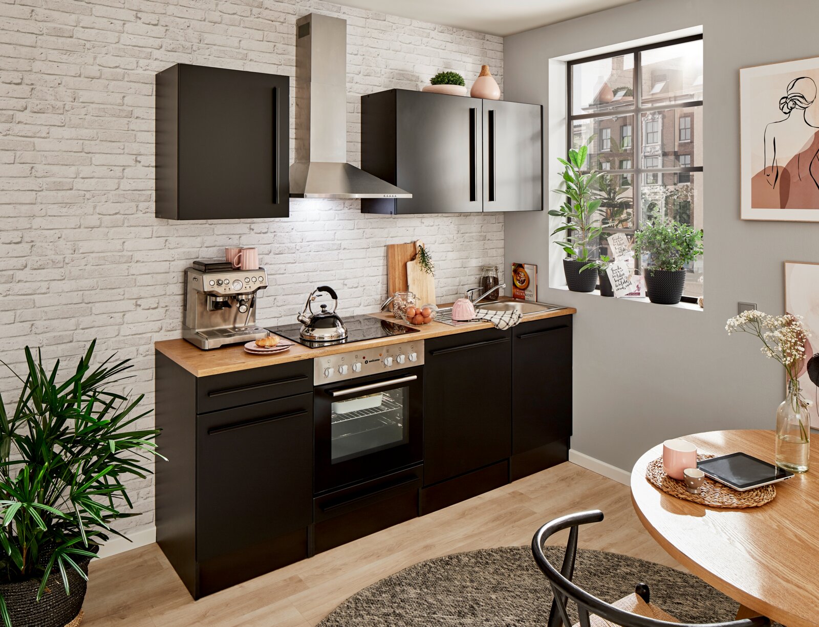 Yourkitchen Keukenblok 220 ZONDER elektrische apparaten JAZZ Een moderne keuken in een zwart ontwerp met een houten werkblad, zonder elektrische apparaten, gezien vanuit een lichte, gezellige ruimte met planten en decoraties.