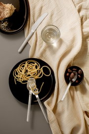 Bovenaanzicht van een minimalistisch gedekte tafel met zwarte borden, roestvrijstalen bestek, een glas water, spaghetti en een schaaltje olijven op een crèmekleurig linnen doek