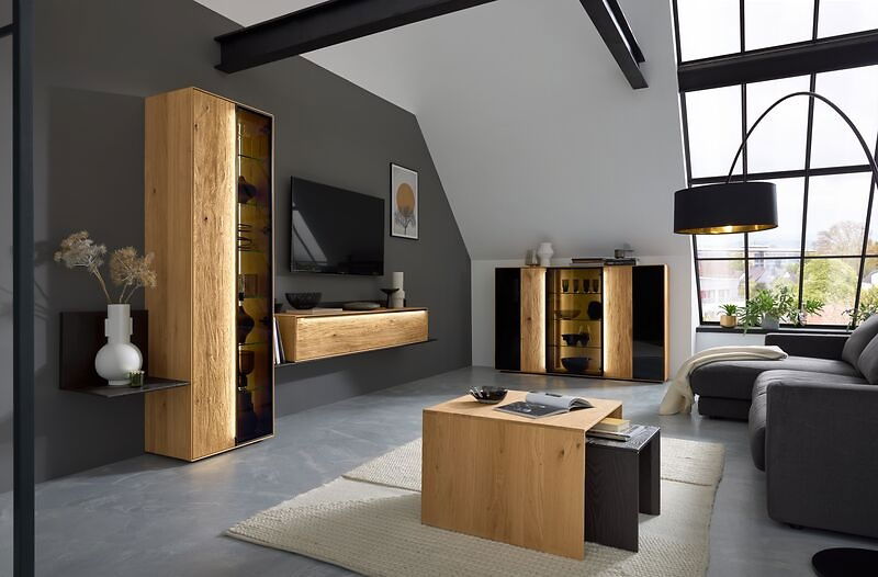 Moderne Wohnzimmeransicht mit einer Wandkombination aus Holz und Glas, einem Beistelltisch und einem Sideboard, beleuchtet durch LED-Leisten, in einem Raum mit großen Fenstern und einem grauen Sofa.