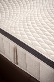 Nahaufnahme einer Matratze mit gestepptem Oberflächenmuster und seitlichem Belüftungsstreifen, perspektivisch von der Seite aufgenommen.