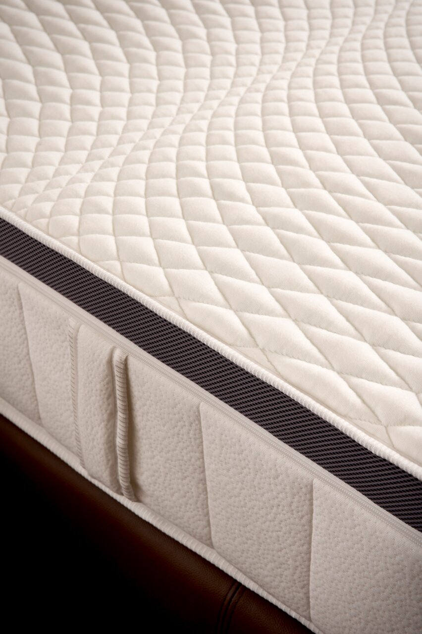 Nahaufnahme einer Matratze mit gestepptem Oberflächenmuster und seitlichem Belüftungsstreifen, perspektivisch von der Seite aufgenommen.