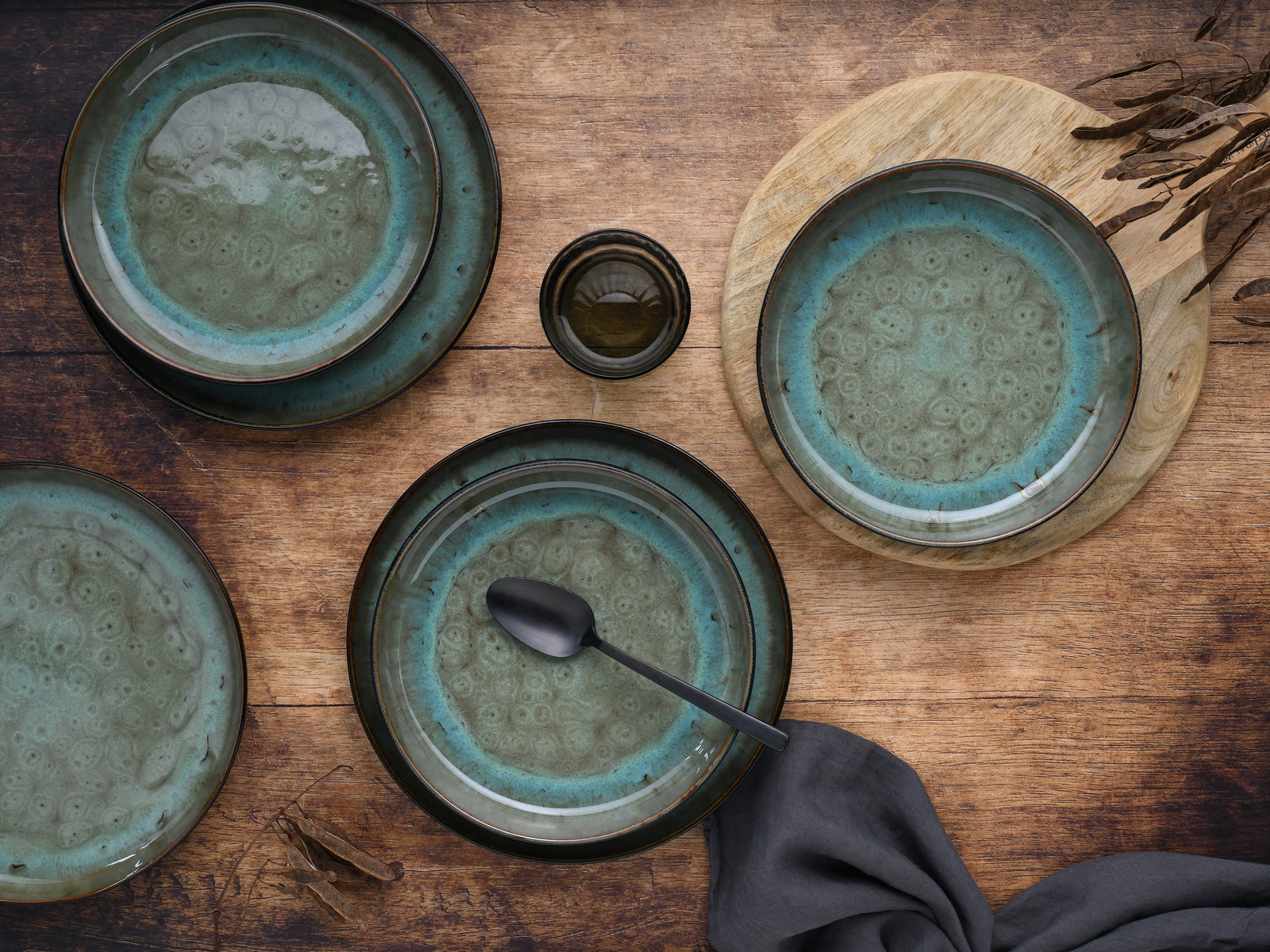 Vogelperspektive auf ein 12-teiliges Tafelservice mit grün-blauen Tellern und Schalen auf einem Holztisch, dekoriert mit einem schwarzen Löffel und einem grauen Tuch.