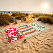 Sommerliches Strand-Setup mit drei gemusterten Strandtüchern (rot-weiß, bunt gestreift, grün-weiß), Liegestuhl mit gelb-weißem Tuch, Strohtasche und Sandalen in den Dünen am Meer bei Sonnenuntergang, Perspektive: leicht erhöhte Vorderansicht (Weitwinkel)