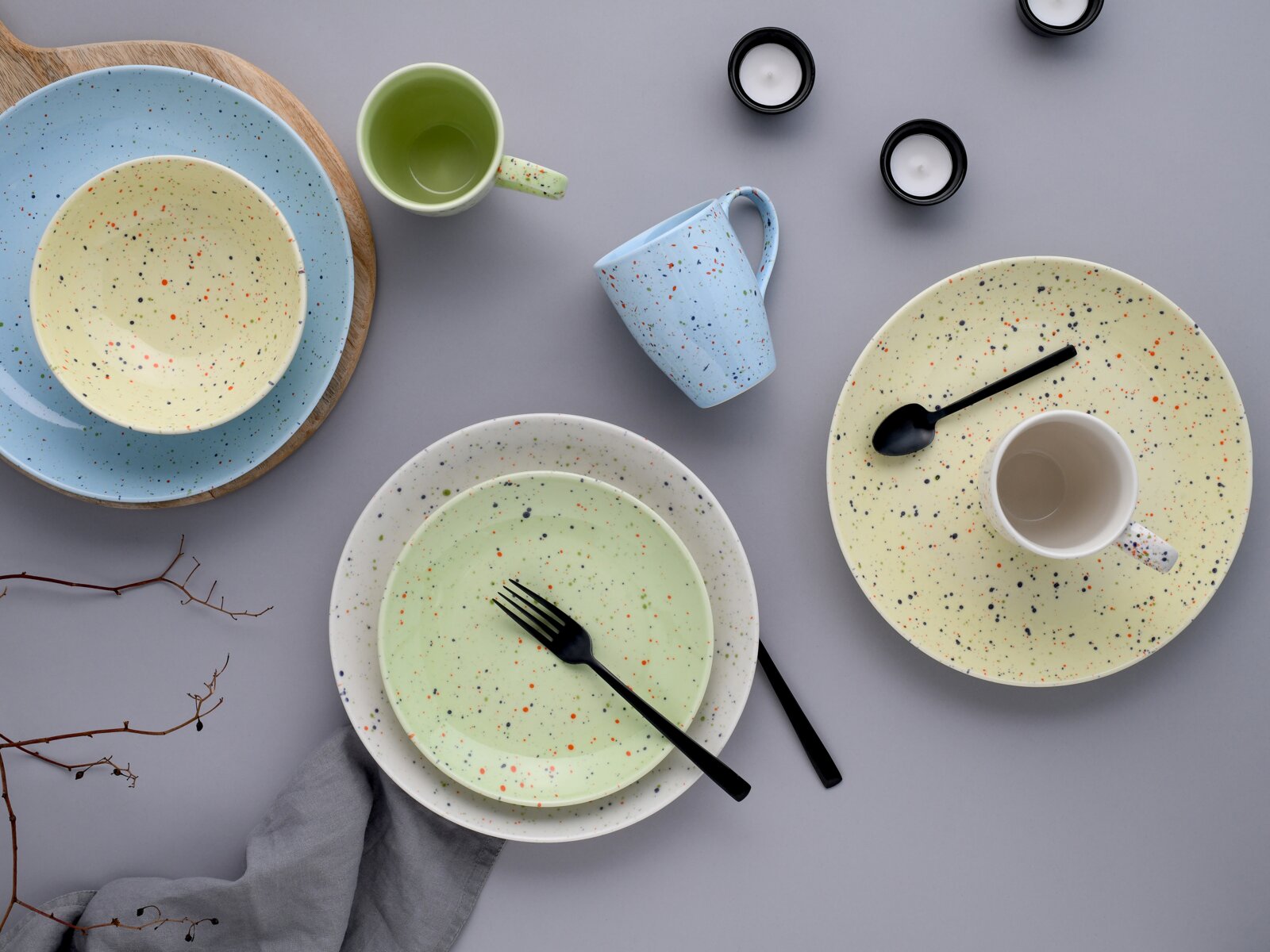 Bunt gesprenkeltes Keramikgeschirr-Set mit Tellern, Schalen und Tassen in Pastellfarben auf grauem Hintergrund, Draufsicht von oben.
