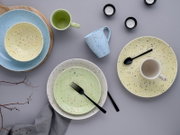 Bunt gesprenkeltes Keramikgeschirr-Set mit Tellern, Schalen und Tassen in Pastellfarben auf grauem Hintergrund, Draufsicht von oben.