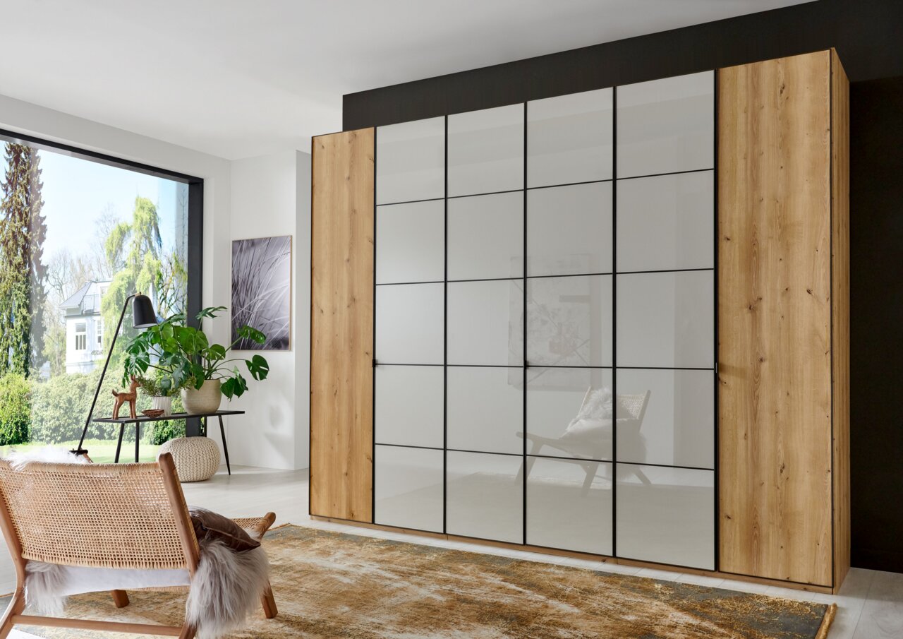 Novel Drehtürenschrank QUITO Moderner Drehtürenschrank mit Holz- und Glaselementen, frontal abgebildet in einem hellen Raum mit großen Fenstern.