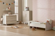Kinderkamerinterieur met beige babybedje, commode en kledingkast, pastelroze wolkenbehang en houten vloer, gefotografeerd in schuin zijaanzicht (breedbeeld) vanaf ooghoogte.