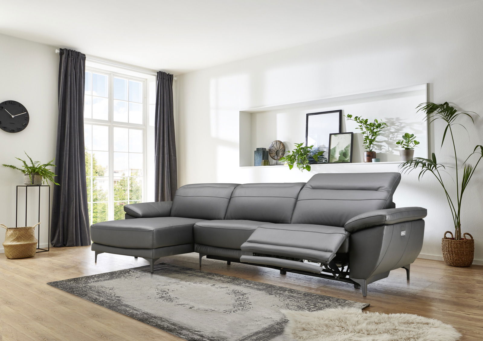 Graue Longchairkombination mit drei Sitzen, rechter Longchair und verstellbarem Fußteil, in einem modernen Wohnzimmer mit großen Fenstern und Pflanzen, seitliche Perspektive.