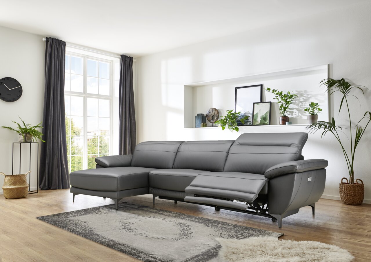Grijze longchair-set met drie zitplaatsen, een longchair aan de rechterkant en een verstelbaar voeteneinde, in een moderne woonkamer met grote ramen en planten, gezien vanaf de zijkant.