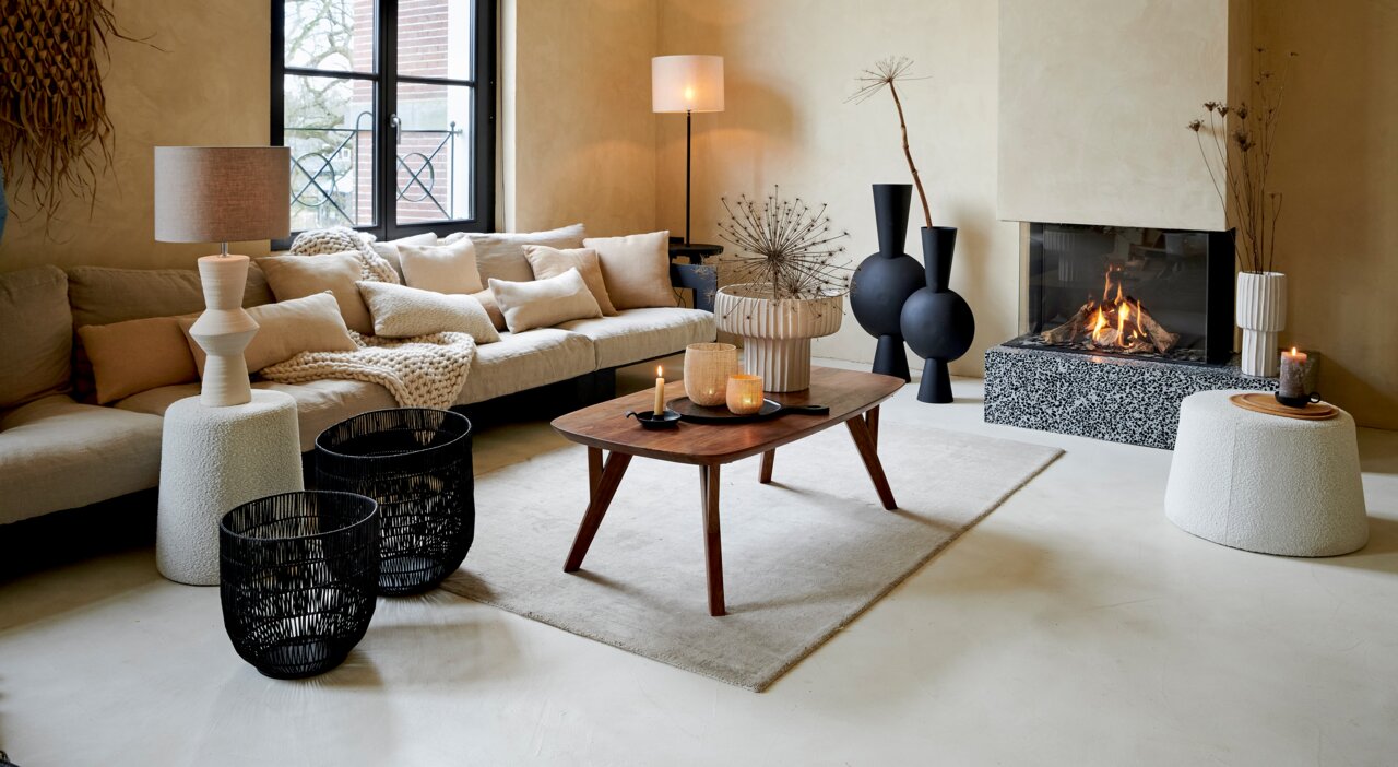 Ein stilvoll eingerichtetes Wohnzimmer mit einem modernen Zylinder-Lampenschirm auf einem Beistelltisch, im Hintergrund ein gemütliches Sofa und ein Kamin. Perspektive: Frontalansicht.