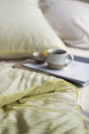 Nahaufnahme einer gestreiften Bettwäsche in sanften Gelbtönen, mit einem Kissen im Hintergrund und einer Tasse auf einem Tablett.