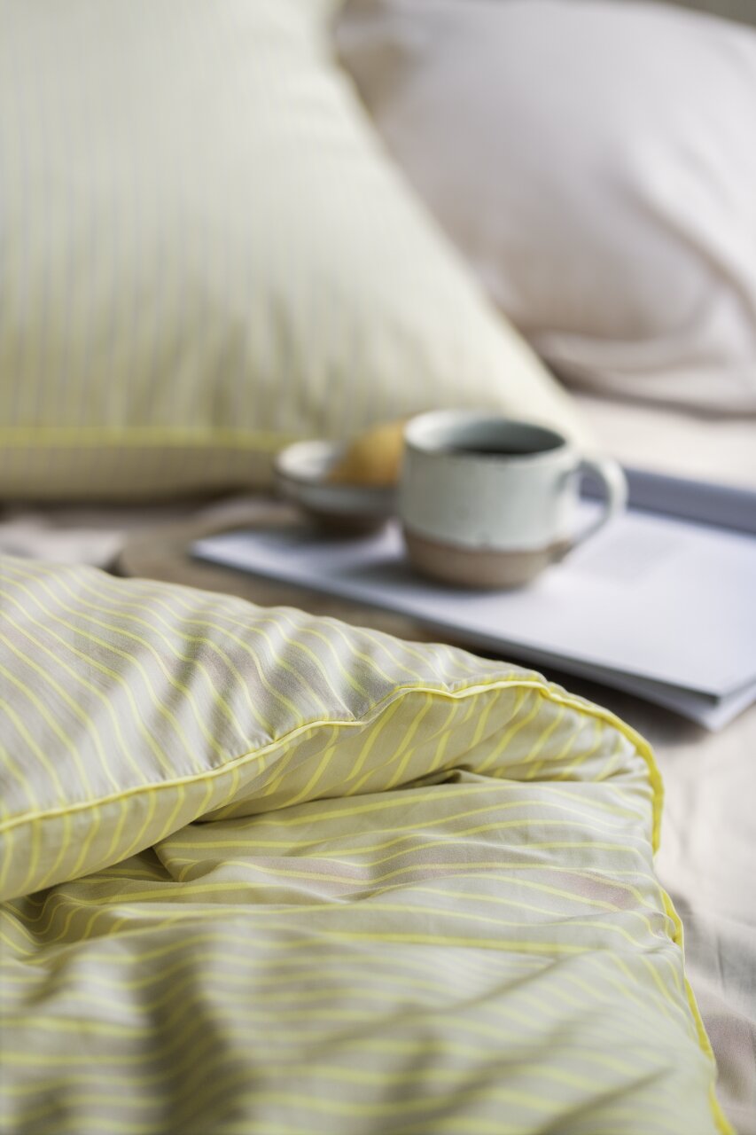 Nahaufnahme einer gestreiften Bettwäsche in sanften Gelbtönen, mit einem Kissen im Hintergrund und einer Tasse auf einem Tablett.