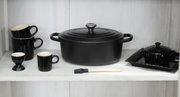 Schwarzer Bräter Ov Sig 29 cm in Frontalansicht, umgeben von schwarzem Geschirr und Küchenutensilien.