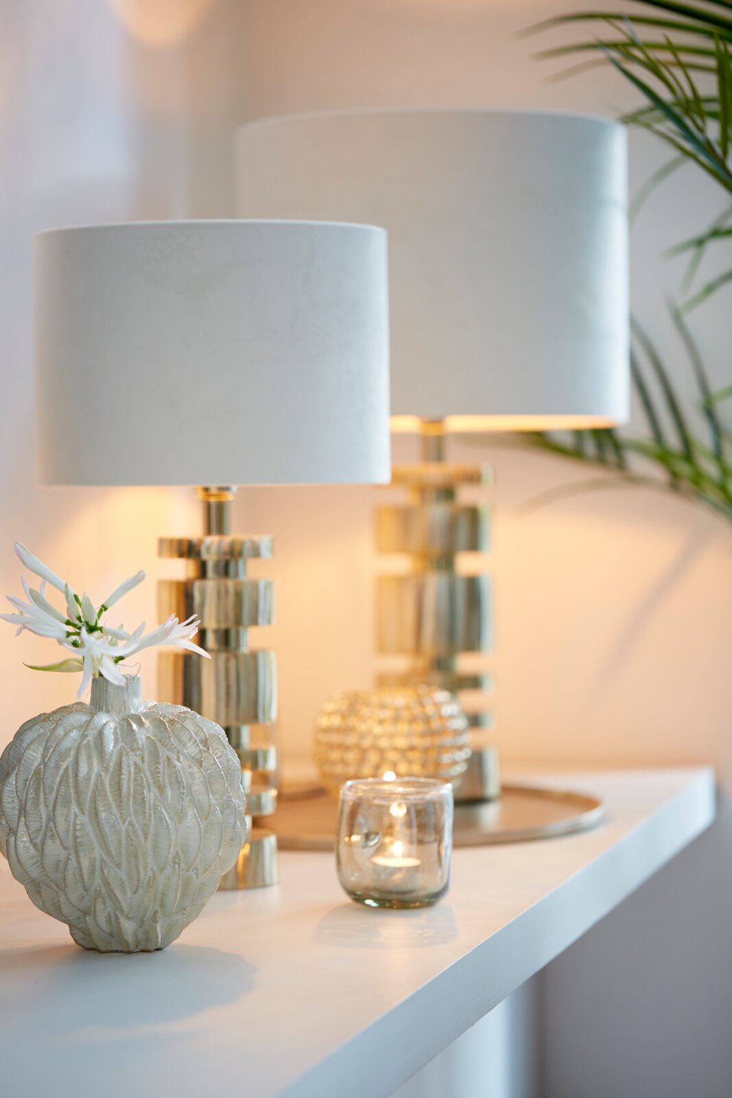 Zwei elegante Tischlampen mit zylindrischen, weißen Lampenschirmen auf einem modernen, goldenen Sockel, seitliche Perspektive.