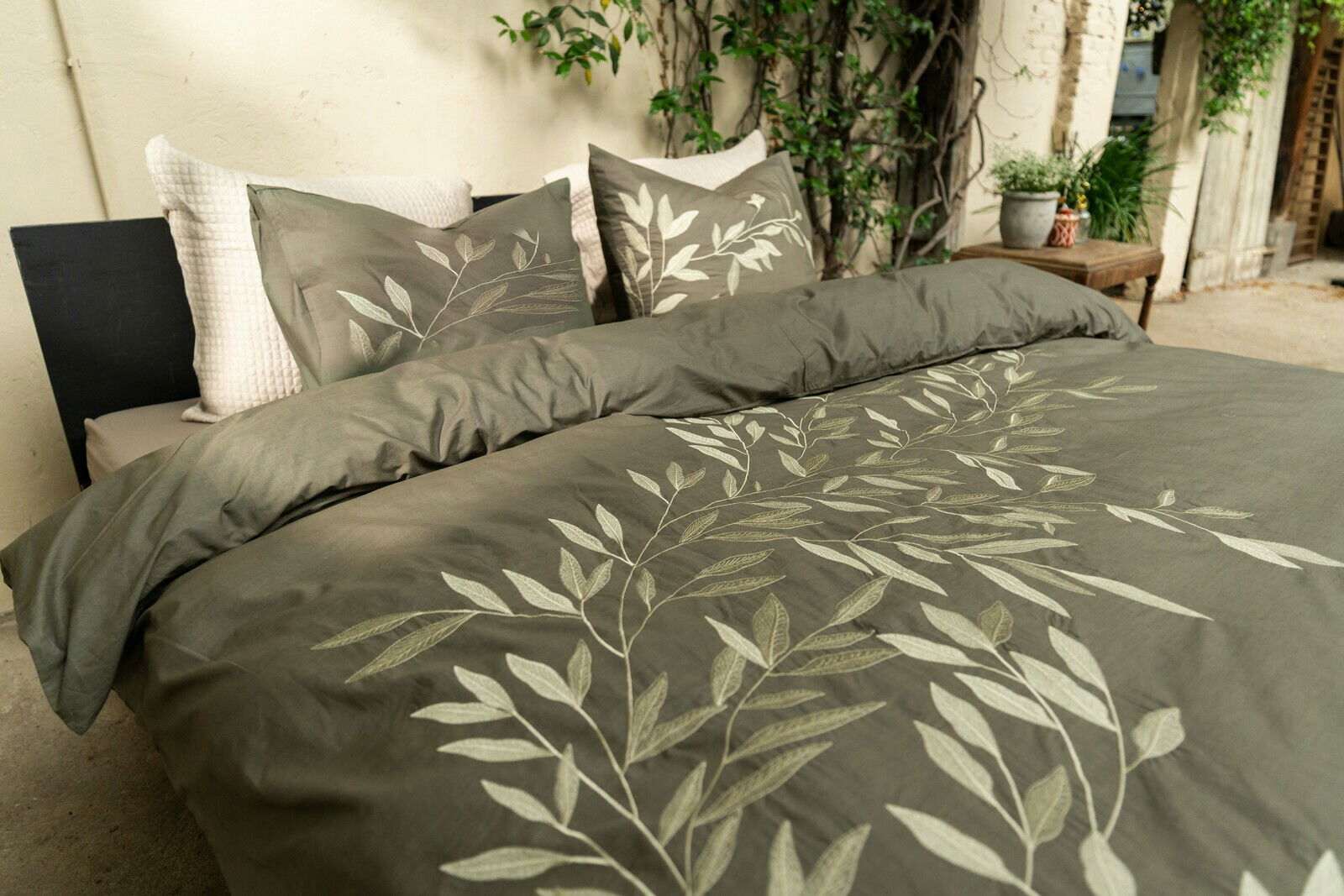 Bettwäsche-Set in einem natürlichen Grünton mit Blattmuster, auf einem Bett arrangiert, umgeben von Pflanzen, aus einer seitlichen Perspektive fotografiert.