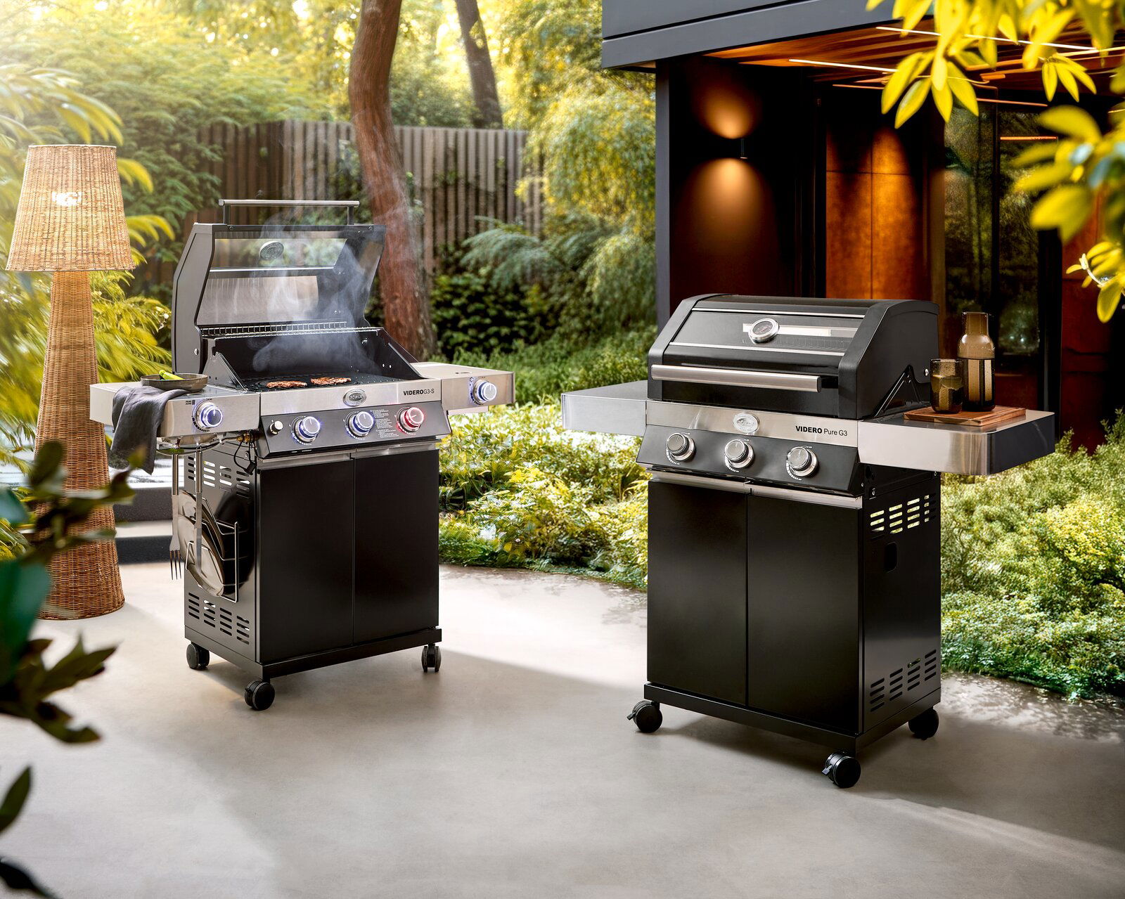 Zwei moderne Gasgrills auf einer Terrasse im Garten, links mit geöffnetem Deckel und sichtbarem Grillgut, rechts mit geschlossenem Deckel und Seitenablage; Perspektive: frontale leicht seitliche Ansicht auf Augenhöhe.