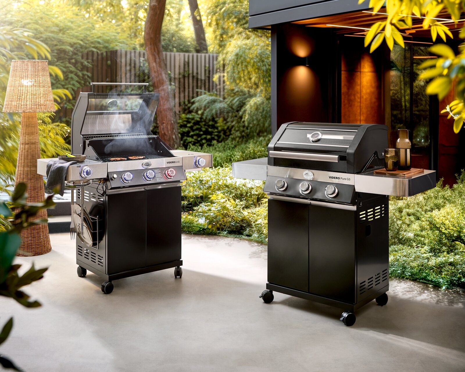 Zwei moderne Gasgrills auf einer Terrasse im Garten, links mit geöffnetem Deckel und sichtbarem Grillgut, rechts mit geschlossenem Deckel und Seitenablage; Perspektive: frontale leicht seitliche Ansicht auf Augenhöhe.