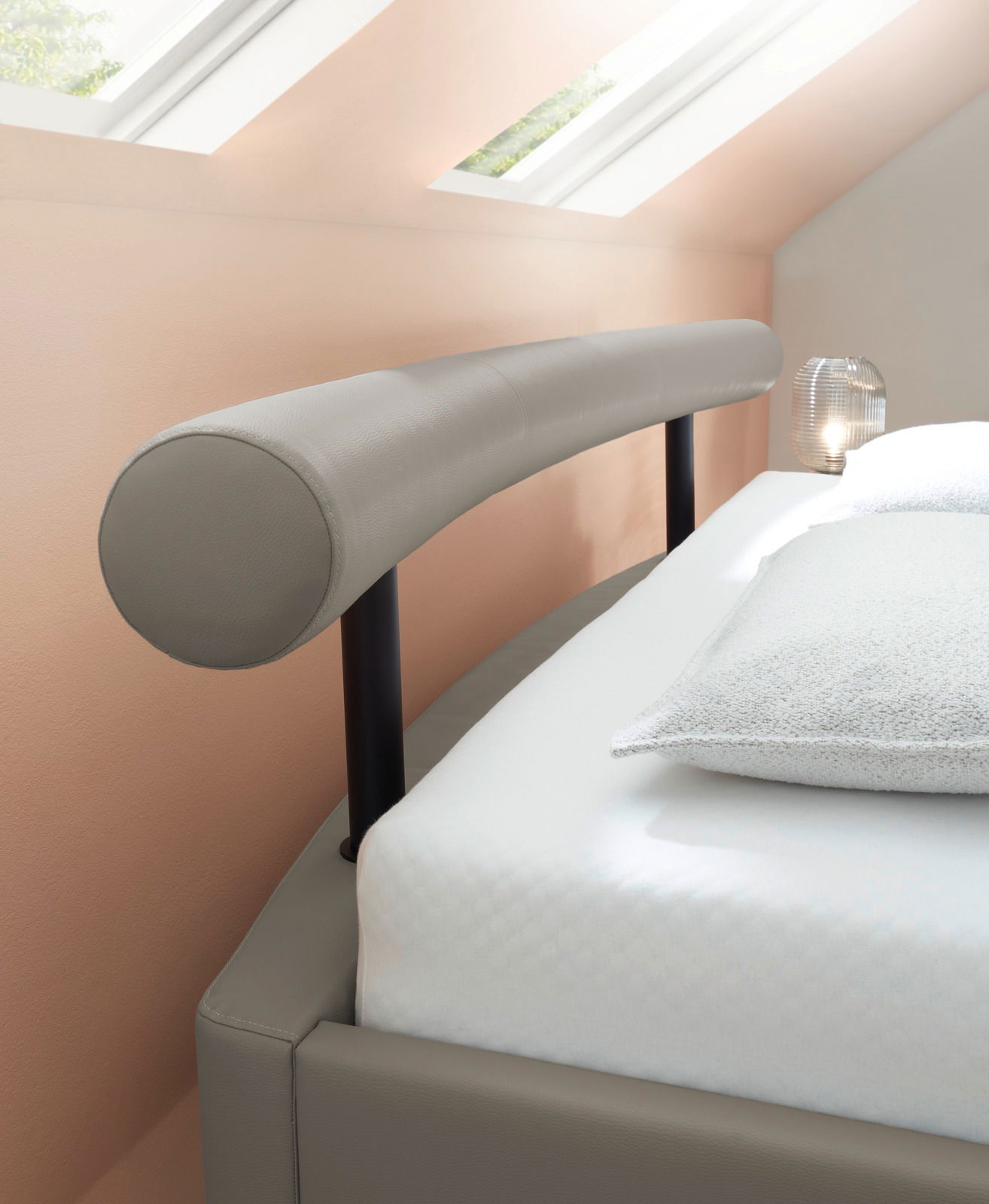 Seitliche Perspektive eines modernen Polsterbetts mit gepolstertem Kopfteil und elegantem Design unter einem Dachfenster.