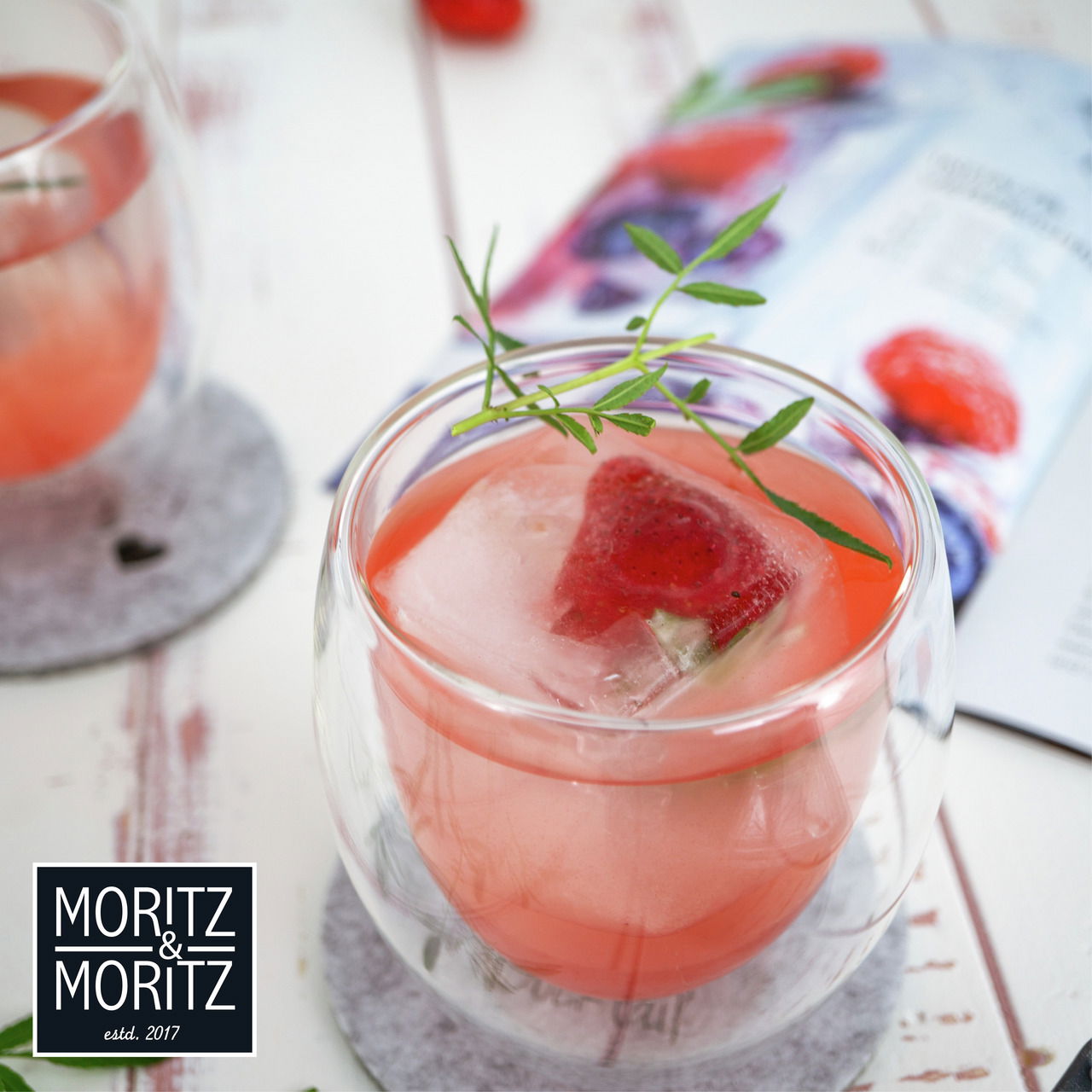 Nahaufnahme eines doppelwandigen Glases mit rosa Getränk und einem Eiswürfel, der eine Erdbeere enthält, auf einem Tisch mit einem Untersetzer.