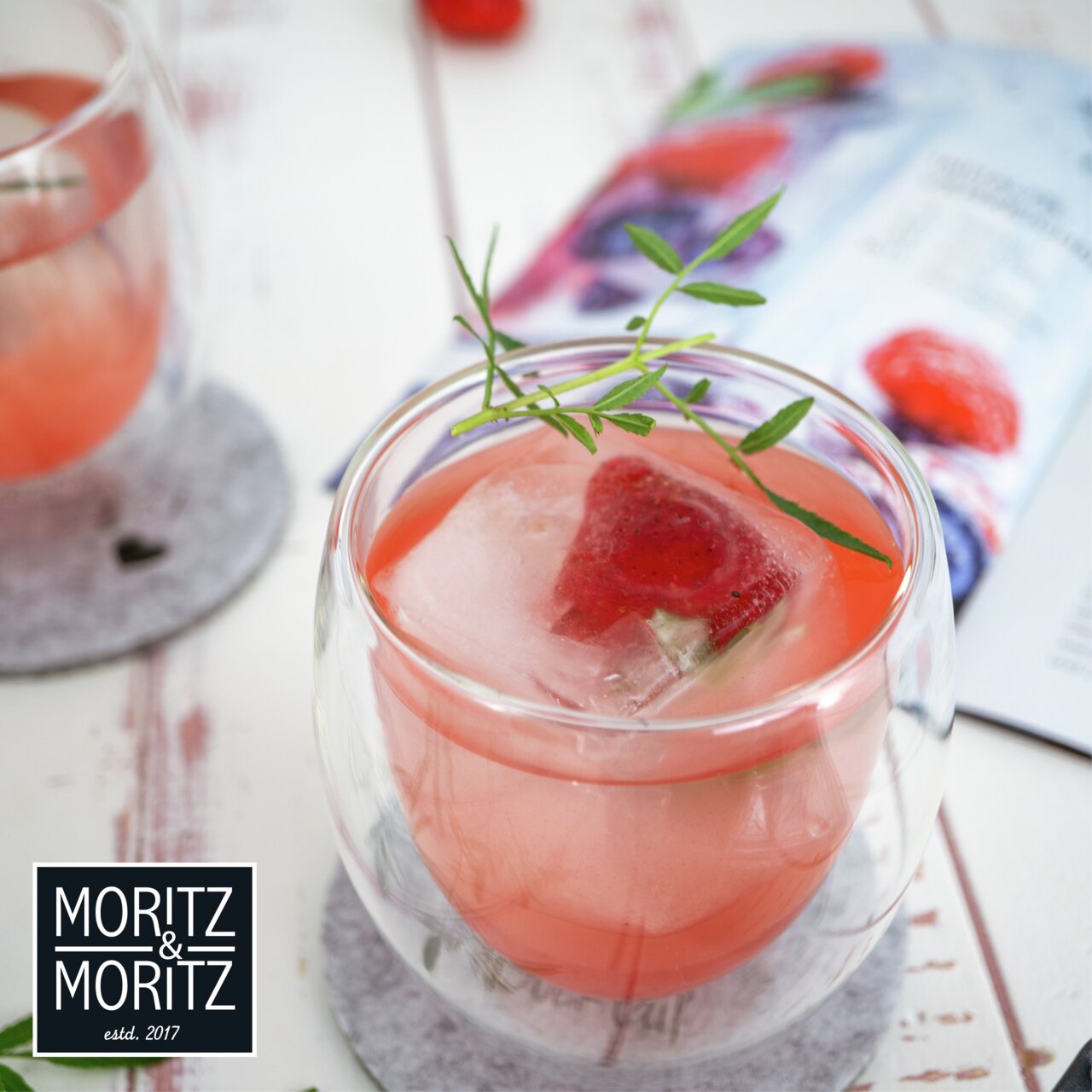 Nahaufnahme eines doppelwandigen Glases mit rosa Getränk und einem Eiswürfel, der eine Erdbeere enthält, auf einem Tisch mit einem Untersetzer.