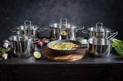 Fünfteiliges Topfset Jaspis aus Edelstahl, frontal fotografiert, mit verschiedenen Zutaten und einer Pfanne mit Gericht im Vordergrund.