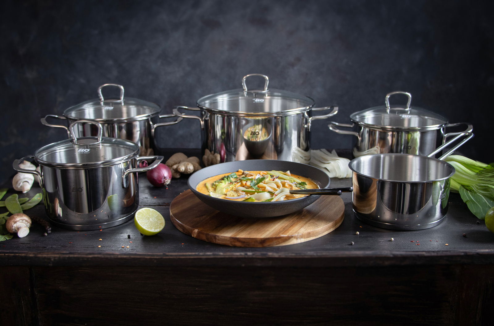 Fünfteiliges Topfset Jaspis aus Edelstahl, frontal fotografiert, mit verschiedenen Zutaten und einer Pfanne mit Gericht im Vordergrund.