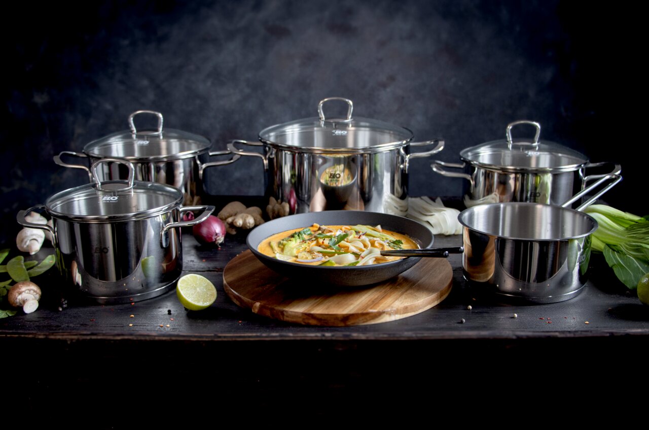 Fünfteiliges Topfset Jaspis aus Edelstahl, frontal fotografiert, mit verschiedenen Zutaten und einer Pfanne mit Gericht im Vordergrund.
