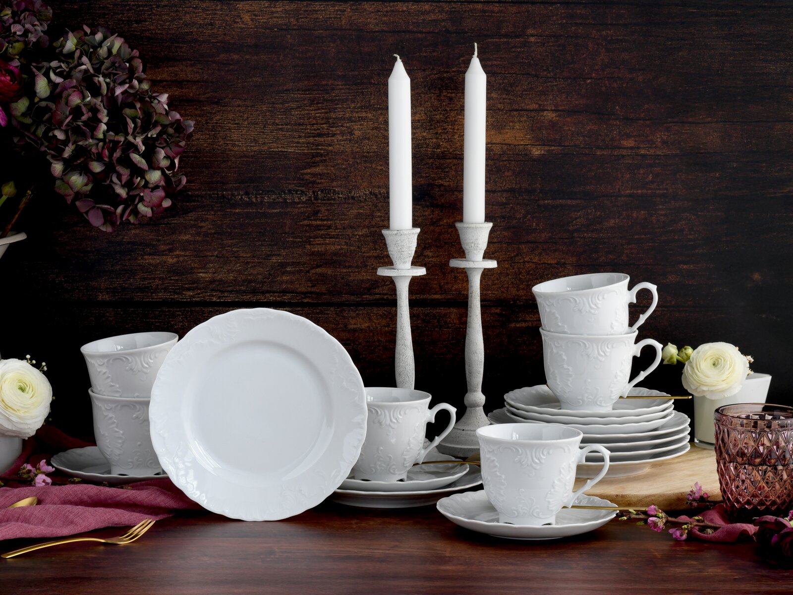 creaTable Kaffeeservice 18-tlg ROKOKO Weißes Porzellan-Geschirrset mit reliefiertem Dekor, Tassen und Tellern auf rustikalem Holztisch, dekoriert mit Kerzenständern und Blumen, fotografiert aus frontaler Perspektive auf Augenhöhe.