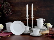 Weißes Porzellan-Geschirrset mit reliefiertem Dekor, Tassen und Tellern auf rustikalem Holztisch, dekoriert mit Kerzenständern und Blumen, fotografiert aus frontaler Perspektive auf Augenhöhe.