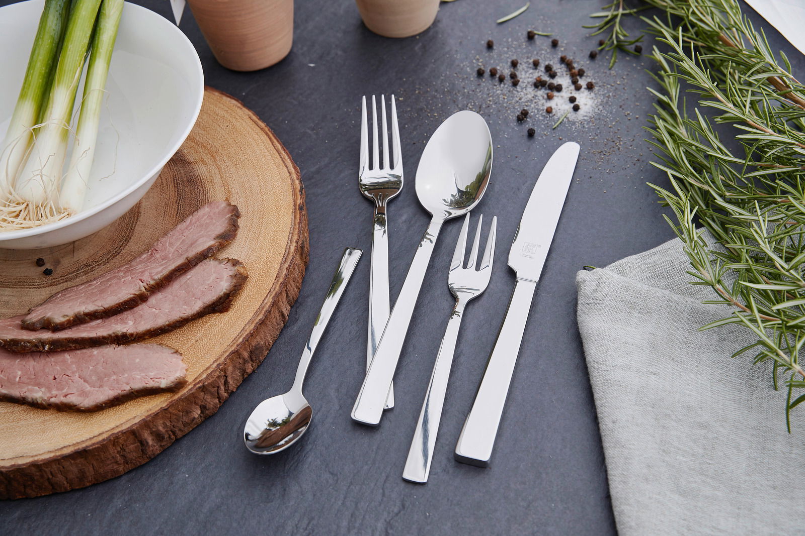 Elegantes Besteckset King 100-tlg. aus Edelstahl, bestehend aus Messer, Gabel, Löffel und Teelöffel, auf einer dunklen Schieferplatte arrangiert, umgeben von frischen Kräutern und Gewürzen. Draufsicht.