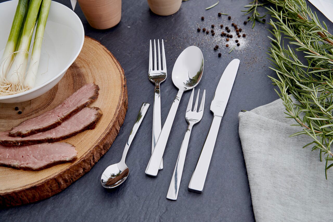 ZWILLING Besteck King 100-tlg. Elegantes Besteckset King 100-tlg. aus Edelstahl, bestehend aus Messer, Gabel, Löffel und Teelöffel, auf einer dunklen Schieferplatte arrangiert, umgeben von frischen Kräutern und Gewürzen. Draufsicht.