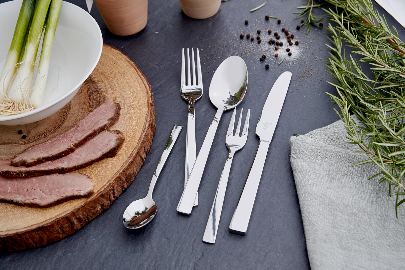 ZWILLING Besteck King 100-tlg. Elegantes Besteckset King 100-tlg. aus Edelstahl, bestehend aus Messer, Gabel, Löffel und Teelöffel, auf einer dunklen Schieferplatte arrangiert, umgeben von frischen Kräutern und Gewürzen. Draufsicht.