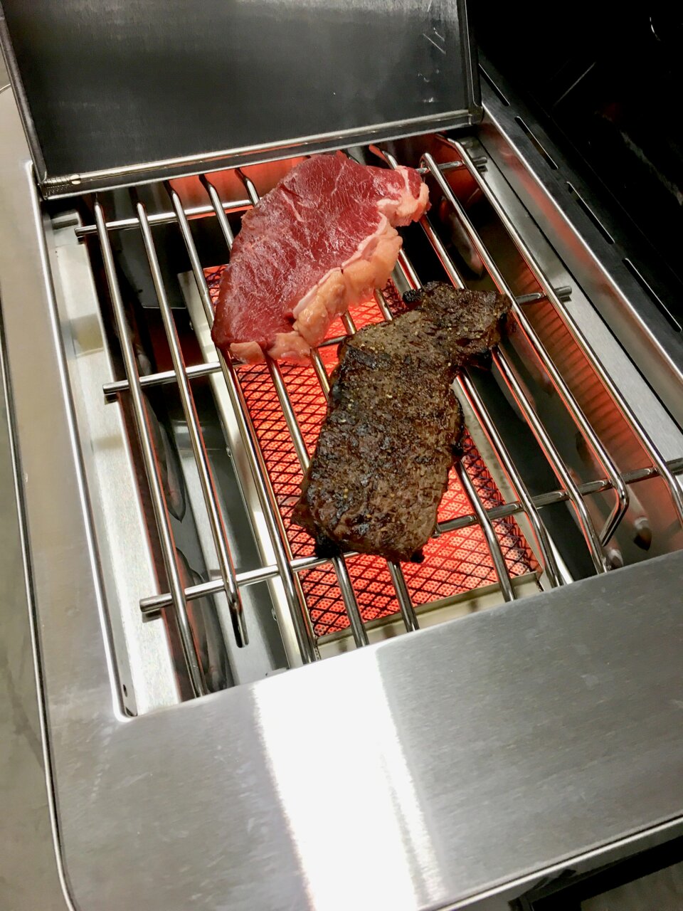 Twee biefstukken op een roestvrijstalen infraroodgrill boven gloeiende keramische stenen, schuin bovenaanzicht