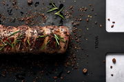 Schwarzes Schneidebrett mit einem gebratenen Fleischstück, garniert mit Rosmarin und Gewürzen, aus der Vogelperspektive.