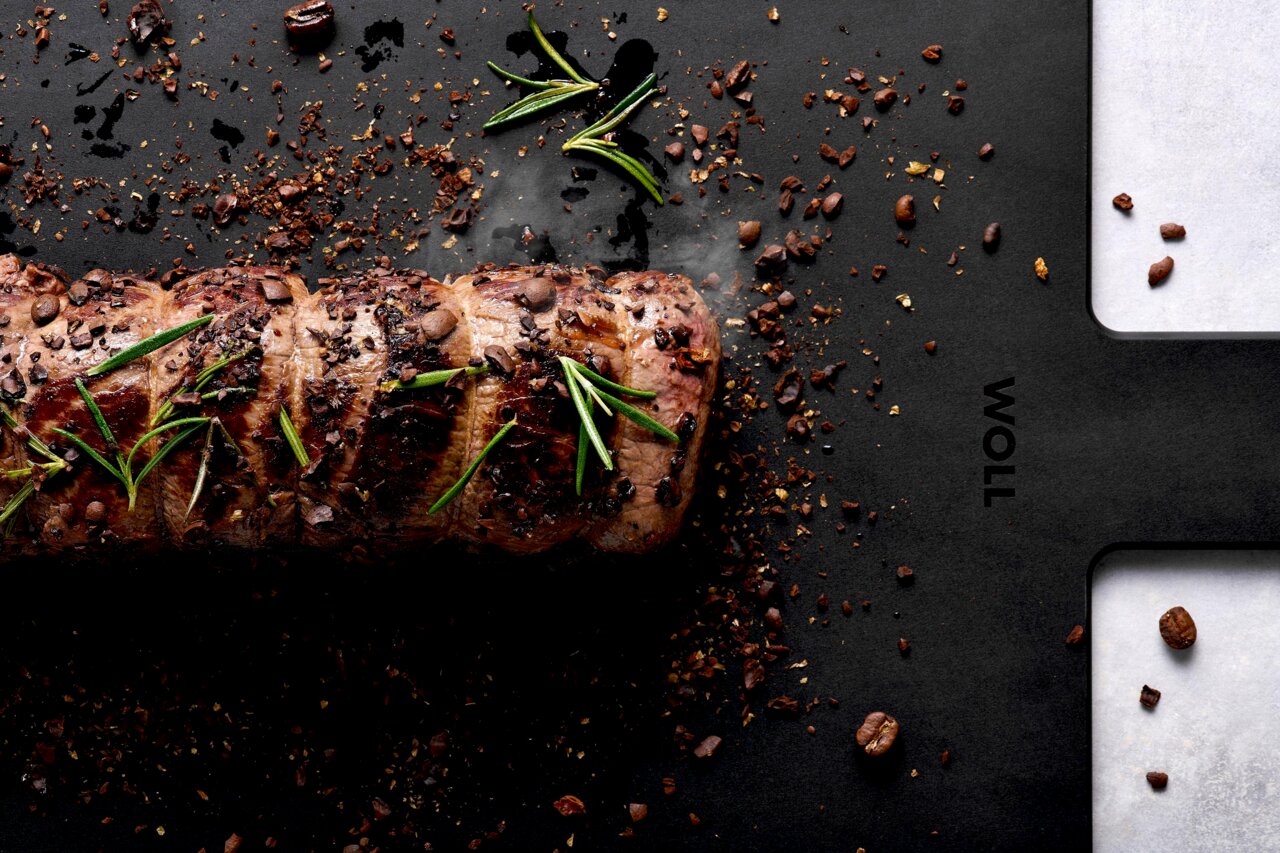 Schwarzes Schneidebrett mit einem gebratenen Fleischstück, garniert mit Rosmarin und Gewürzen, aus der Vogelperspektive.