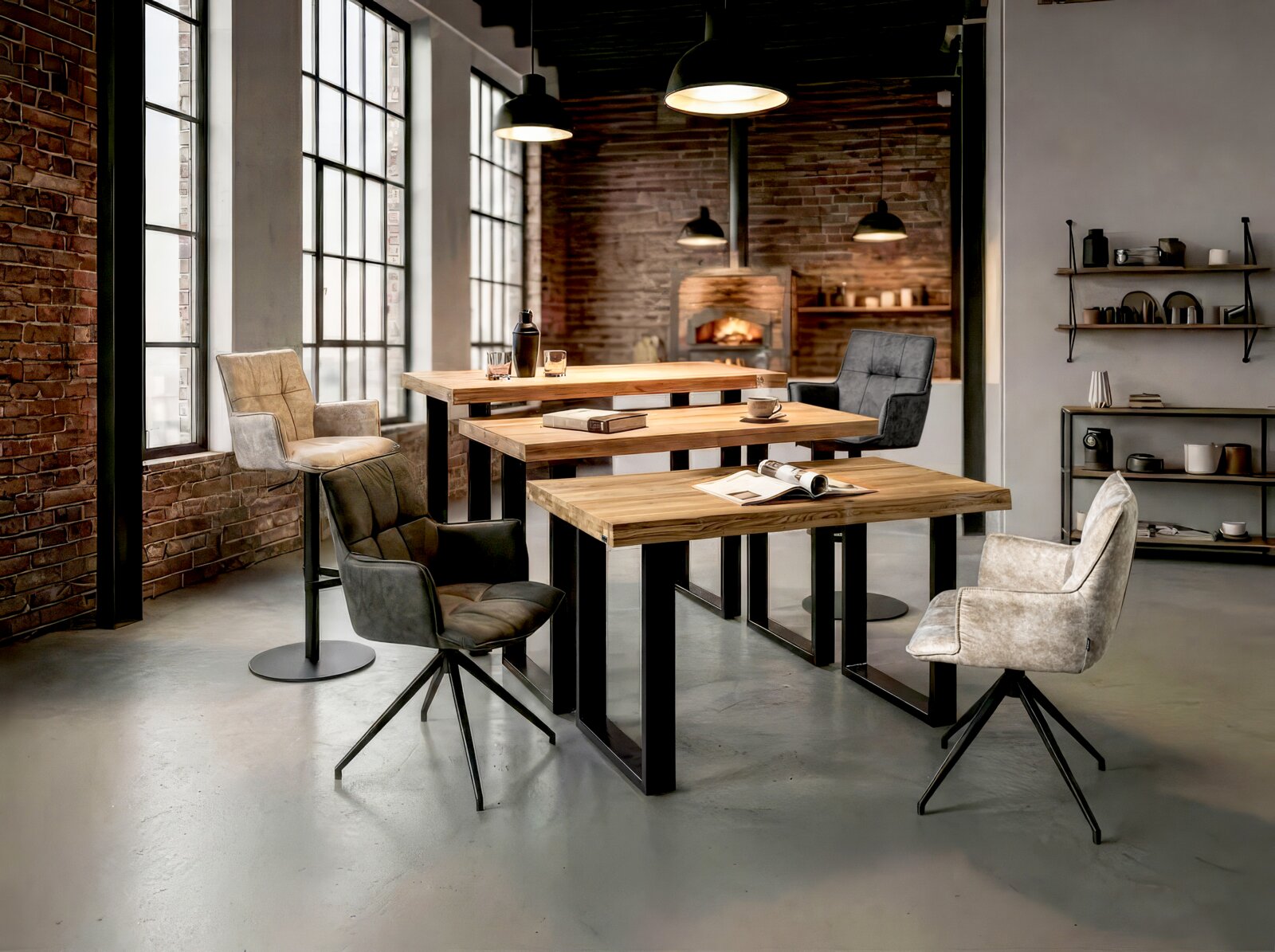 Novel Fauteuil OSAKA Set van drie industriële eettafels met massief houten blad en zwart metalen onderstel, omringd door gestoffeerde stoelen in een loftinterieur met bakstenen muur en grote ramen, gefotografeerd in schuin vooraanzicht op ooghoogte.