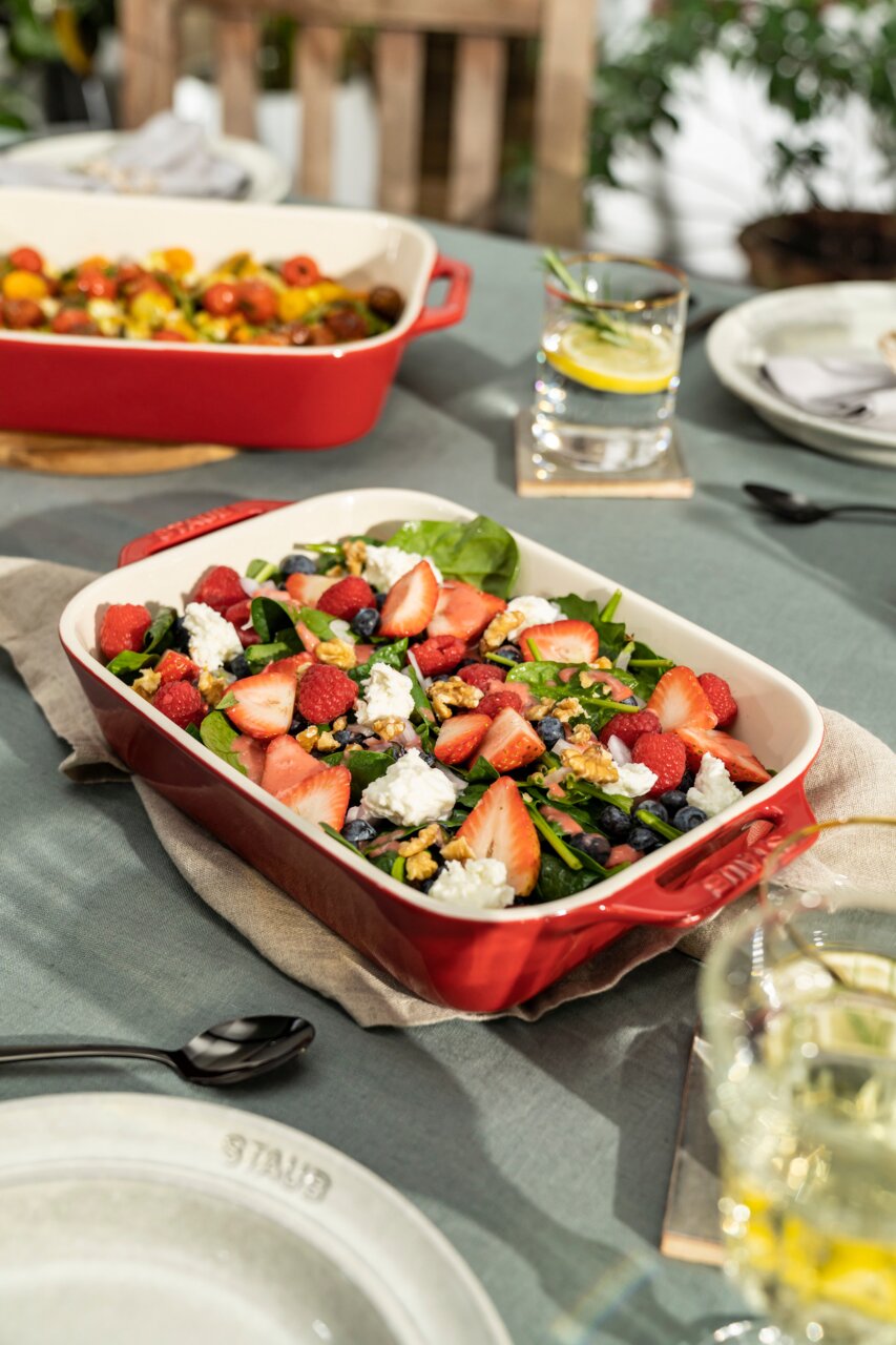 Een rode ovenschaal met een frisse salade van aardbeien, frambozen, bosbessen, walnoten en kaas, van bovenaf gefotografeerd op een gedekte tafel.