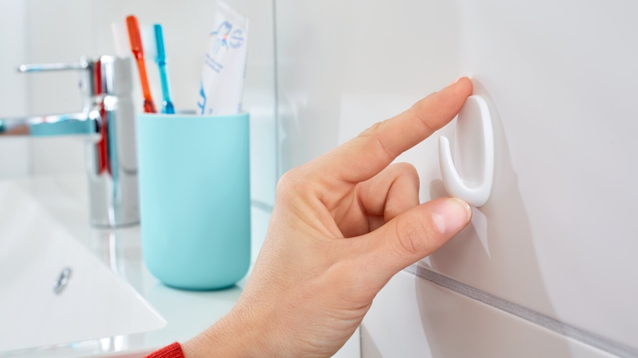 Eine Hand befestigt einen weißen tesa Powerstrips Haken Small an einer Badezimmerfliese. Im Hintergrund sind ein Wasserhahn und ein Zahnputzbecher zu sehen.