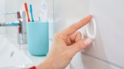 Eine Hand befestigt einen weißen tesa Powerstrips Haken Small an einer Badezimmerfliese. Im Hintergrund sind ein Wasserhahn und ein Zahnputzbecher zu sehen.