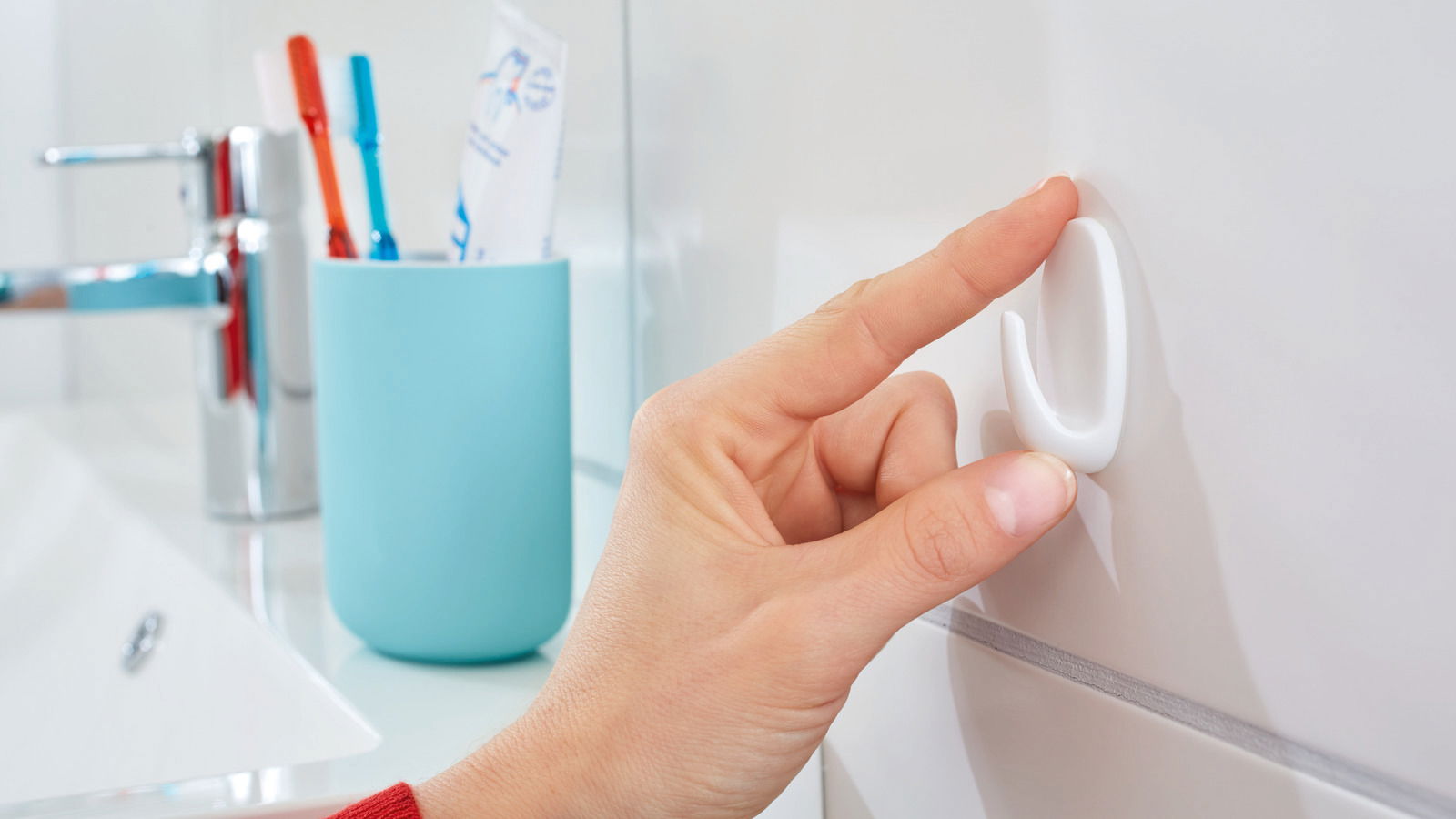Eine Hand befestigt einen weißen tesa Powerstrips Haken Small an einer Badezimmerfliese. Im Hintergrund sind ein Wasserhahn und ein Zahnputzbecher zu sehen.