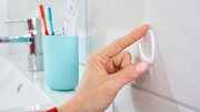 Een hand bevestigt een witte tesa Powerstrips-haak (Small) aan een badkamertegel. Op de achtergrond zijn een kraan en een tandenborstelbeker te zien.