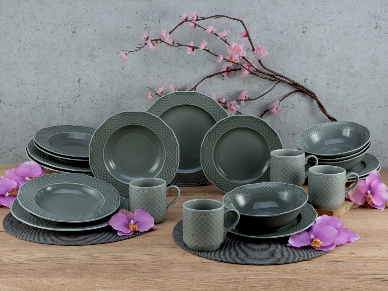 Graues Keramik-Geschirrset mit strukturiertem Rand (Teller, Schalen und Becher) auf Holztisch vor Betonwand, dekoriert mit rosa Blütenzweigen und Orchideen, aufgenommen in frontaler leicht erhöhter Perspektive.