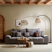 Großes graues Sofa TAVANI in einem gemütlichen Wohnzimmer, frontal fotografiert, mit mehreren Kissen und einem kleinen Hund darauf.