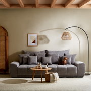 Großes graues Sofa TAVANI in einem gemütlichen Wohnzimmer, frontal fotografiert, mit mehreren Kissen und einem kleinen Hund darauf.