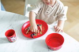 Kind isst von einem roten Kinderteller mit Bauernhofmotiv, begleitet von einer roten Tasse und Schüssel, auf einem Marmortisch aus der Vogelperspektive.