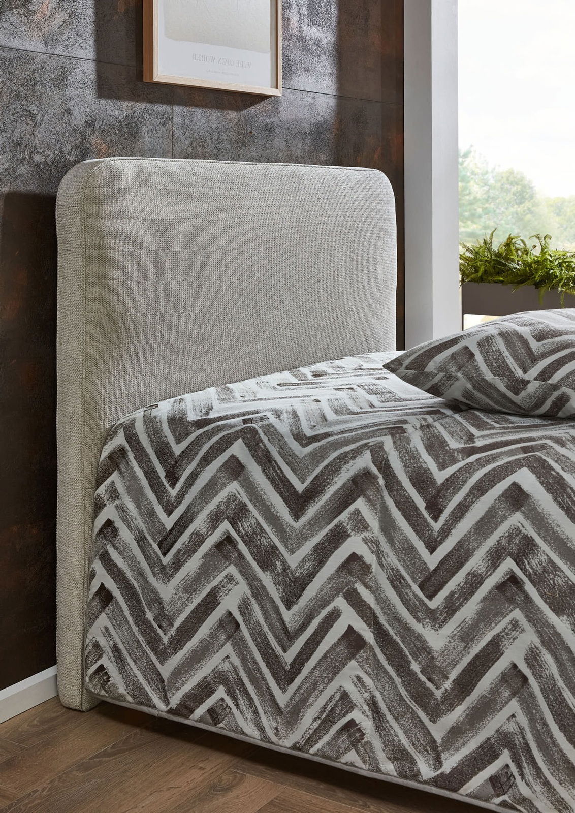 Seitliche Ansicht eines Polsterbetts mit grauem Kopfteil und gemustertem Bettbezug in einem modernen Schlafzimmer.